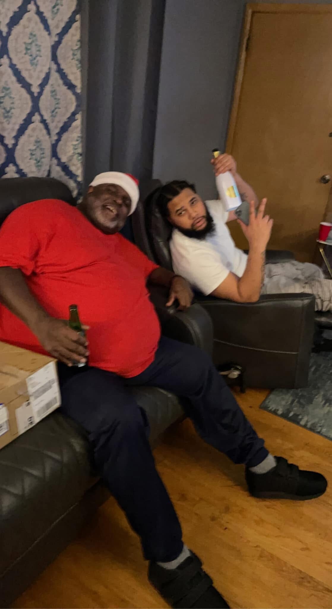 Two men relaxing on a sofa indoors, one in a red shirt and Santa hat holding a beer, the other in a white shirt showing a peace sign with a bottle in hand.