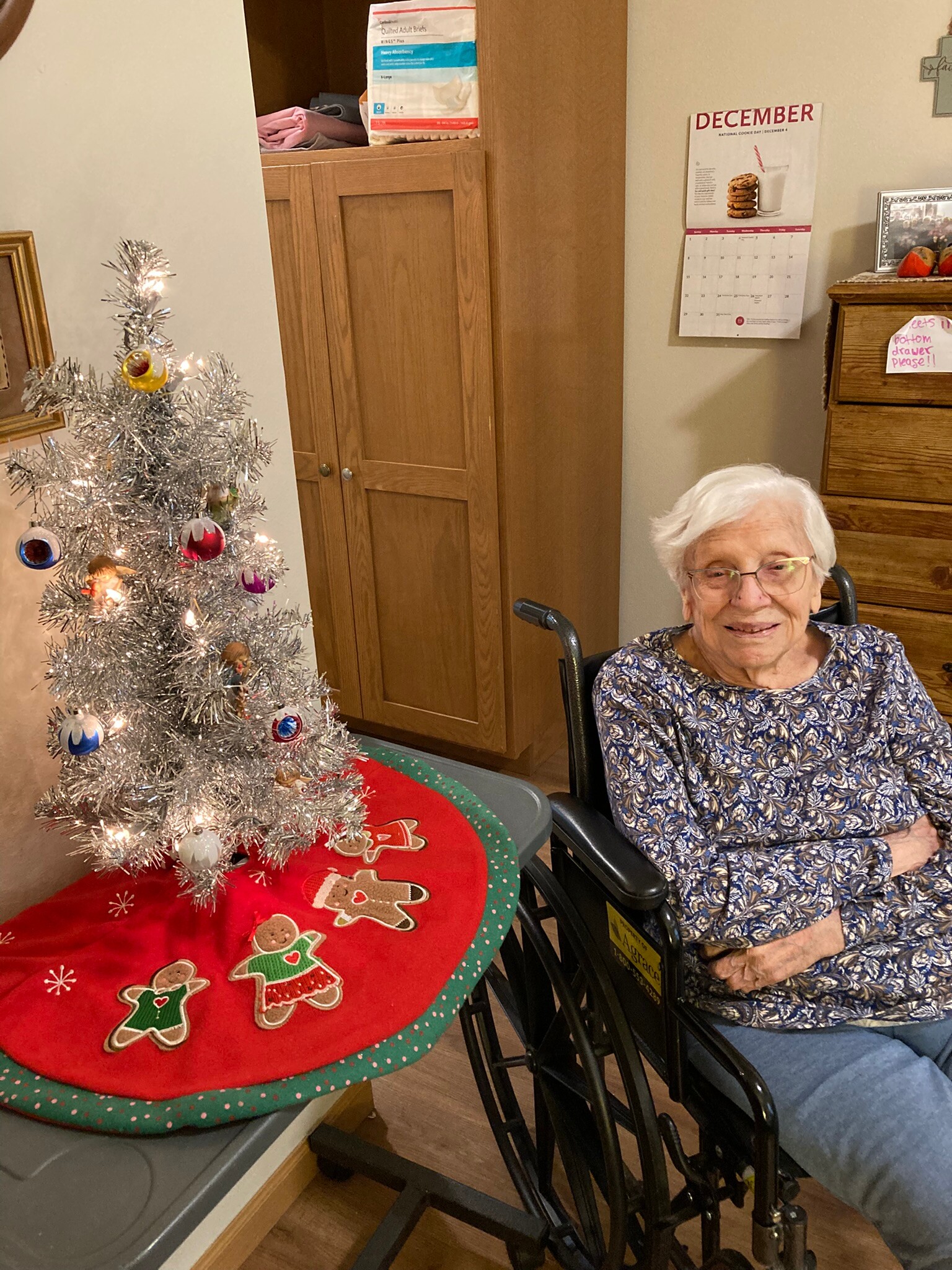 An elderly woman in a wheelchair sits at a Christmas-themed table with a small silver Christmas tree and gingerbread decorations nearby.