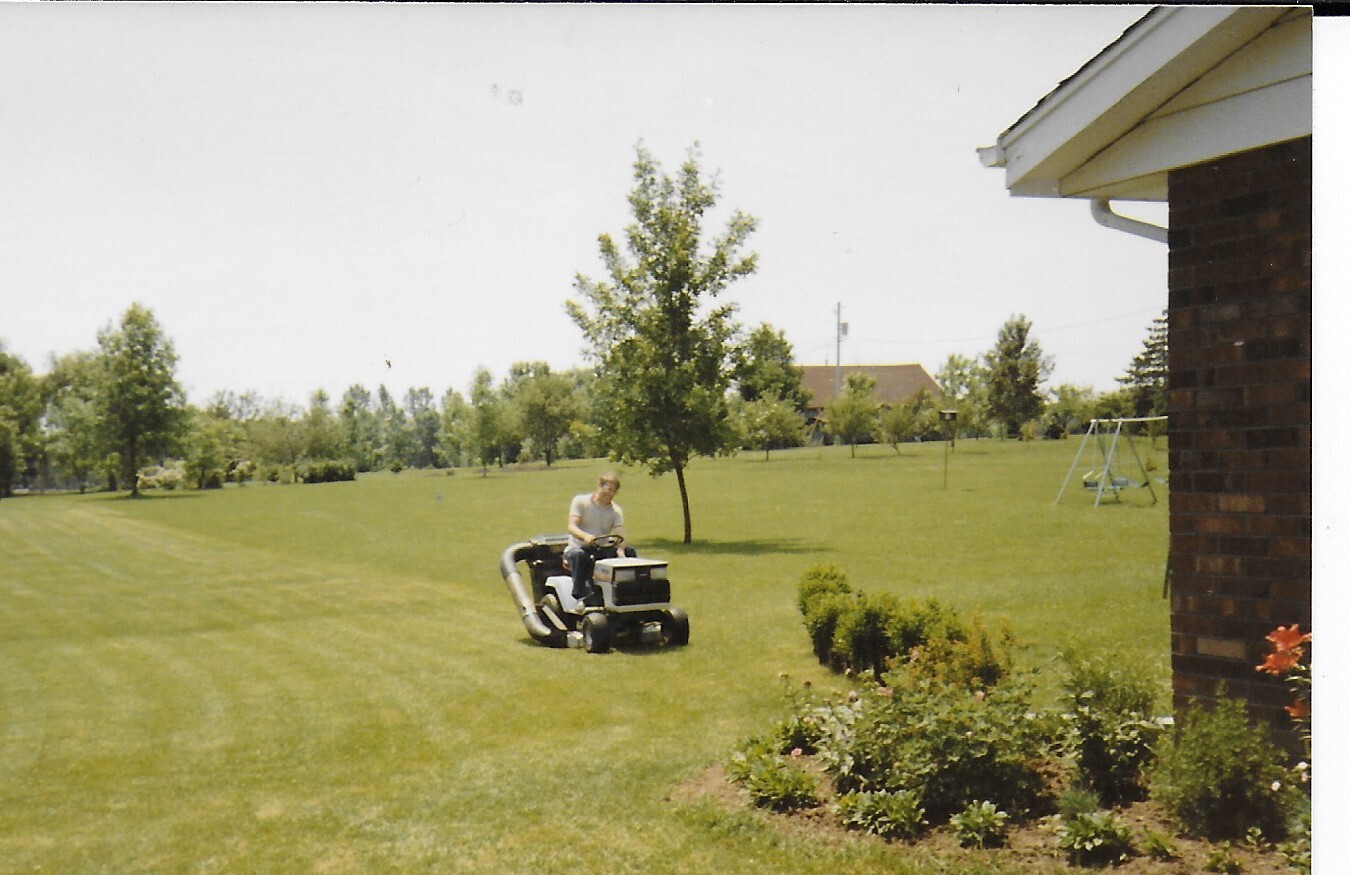 Person riding a lawn mower across a large grassy yard near a brick house.