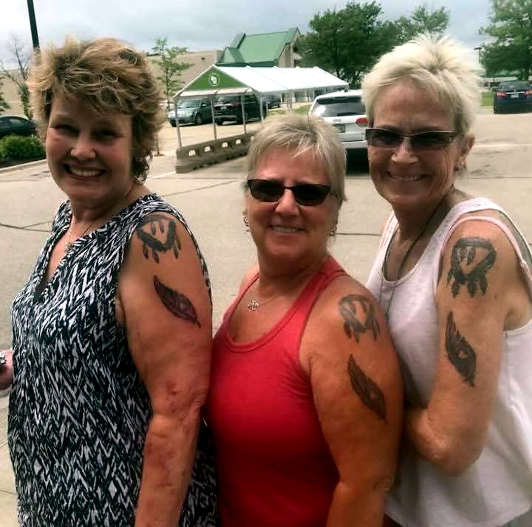 Three smiling women posing together in a parking lot, each with bold feather-shaped tattoo designs on their upper arms.