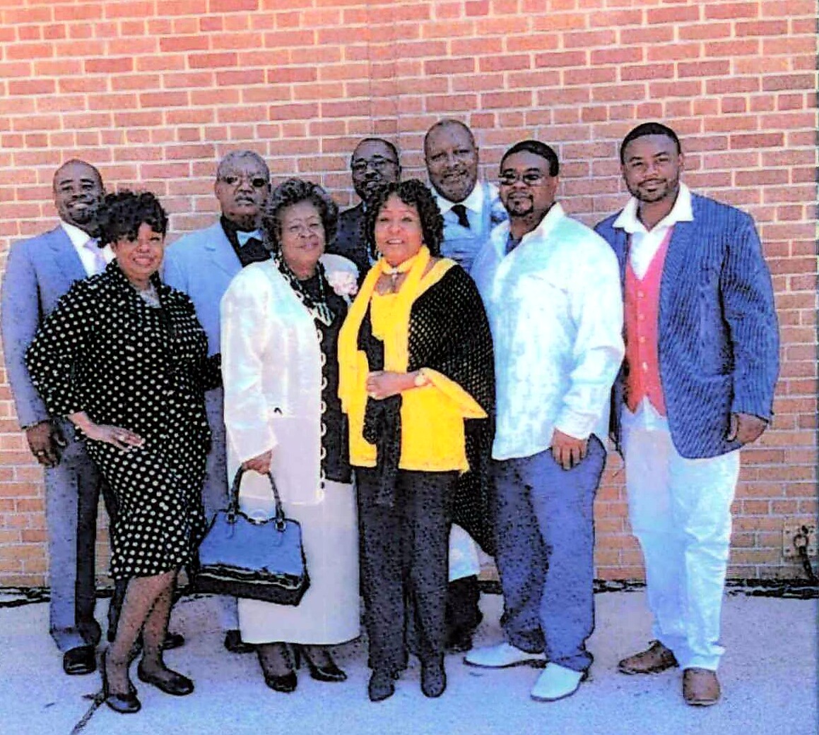 Group of eight adults posing together in front of a brick wall; wearing formal and semi-formal outfits.
