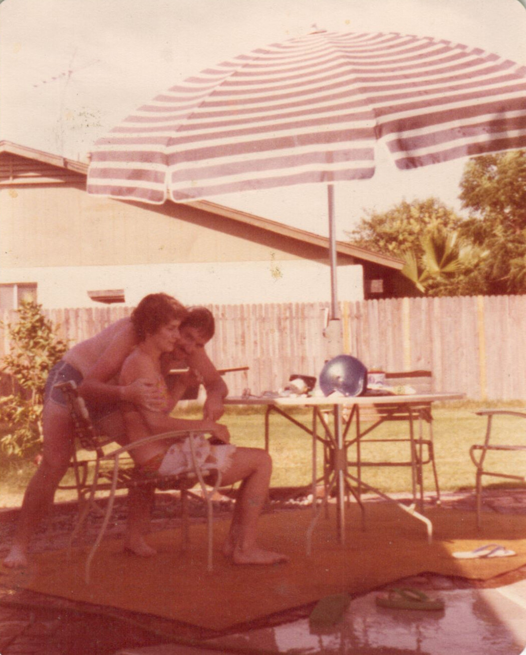Two adults in swimsuits sit together at a metal patio chair in a sunlit backyard, leaning close under a large striped umbrella.