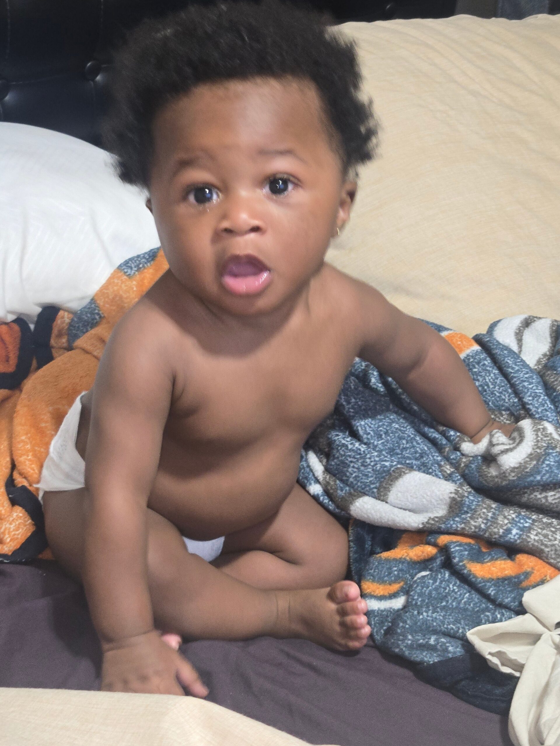 Baby with dark curly hair sits shirtless on a bed, looking surprised with mouth open, wrapped in orange and blue blankets.