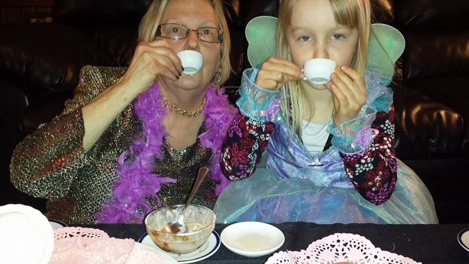 An elderly woman and a young girl in fairy costumes sip tea from tiny cups at a table with doilies.