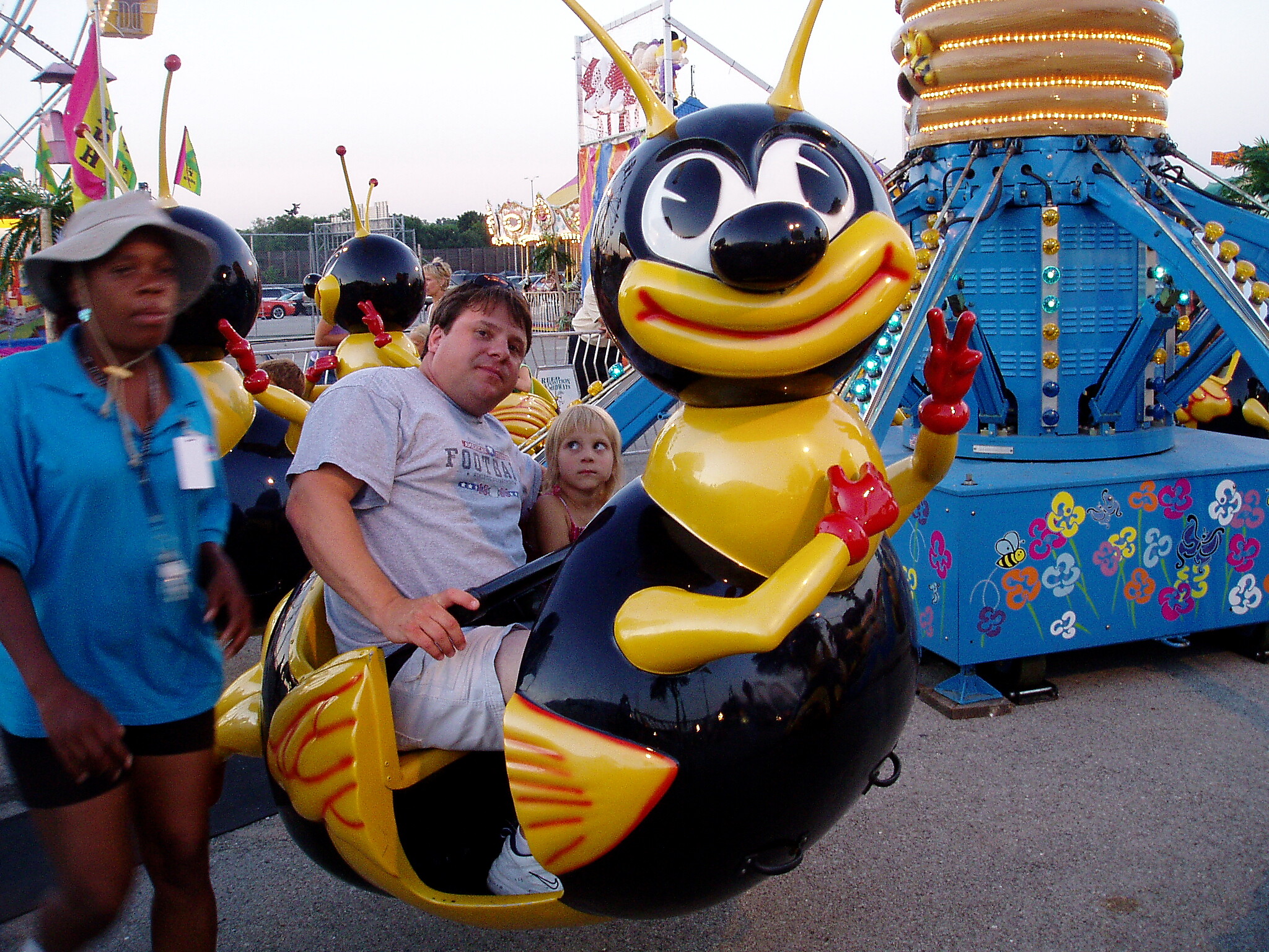 People riding a colorful cartoon-themed amusement ride with a large yellow and black bee character in the foreground, smiling riders nearby.