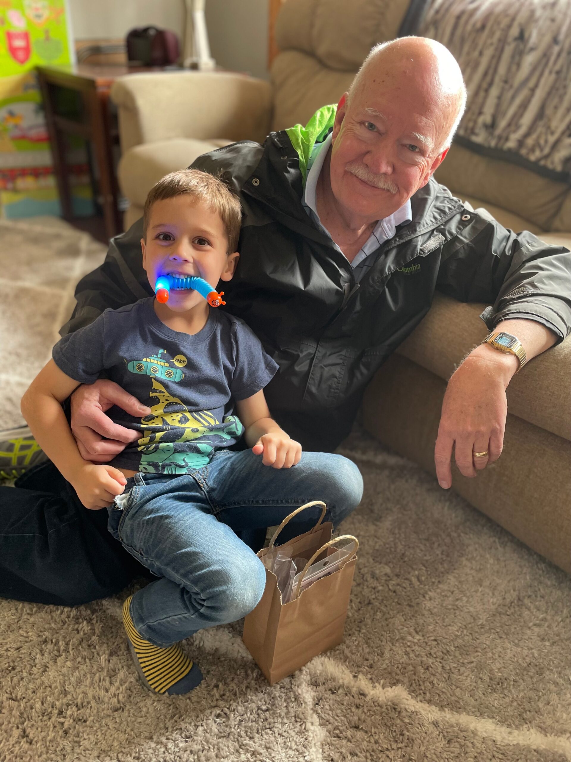 Grandfather and young boy sit together on a beige living room rug; the boy smiles with a blue and orange chew toy in his mouth.