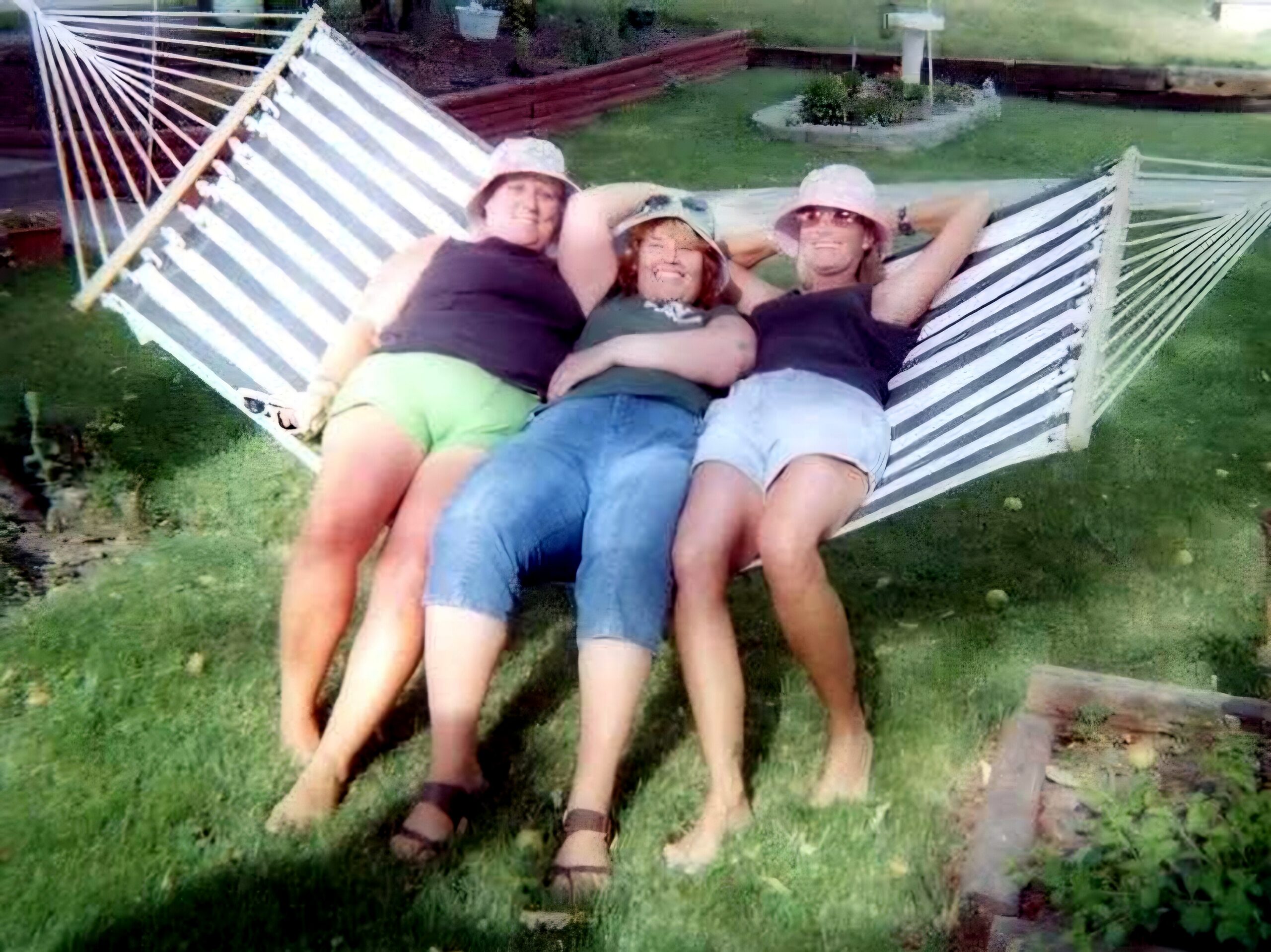 Four friends lounging together in a white-and-gray striped hammock in a backyard, smiling at the camera.