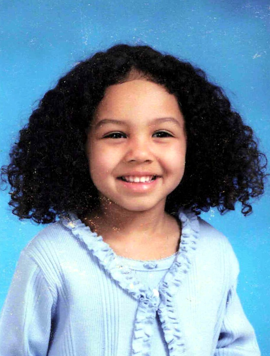 Smiling young girl with dark curly hair wearing a light blue cardigan against a blue background.