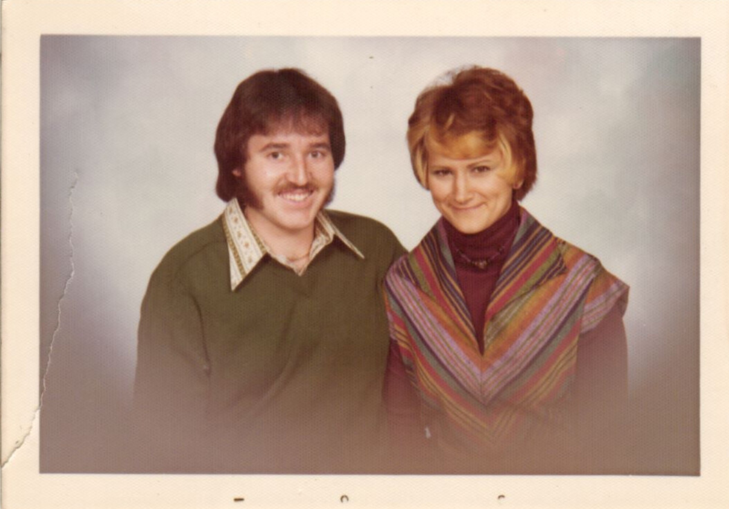 A couple posing together for a portrait, both smiling, with 1970s-style clothing and a soft backdrop.