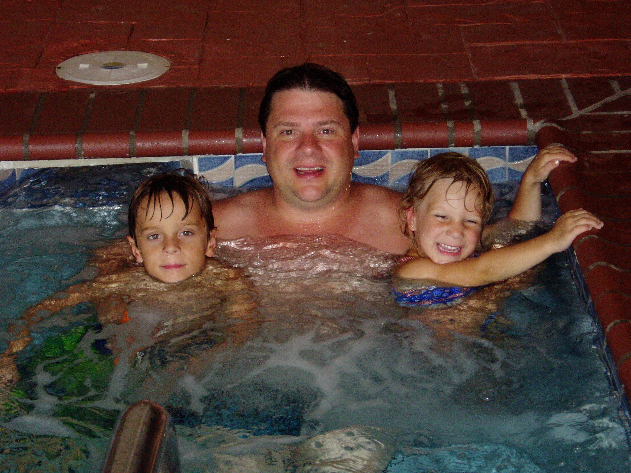 Man and two children smiling in a pool indoors, with a red tiled wall behind them.