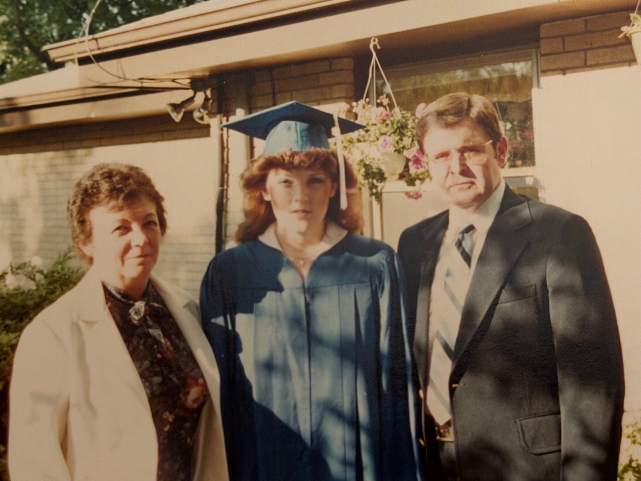 Three adults pose outside a house; a graduate in a blue gown stands between two relatives in formal wear, celebrating.