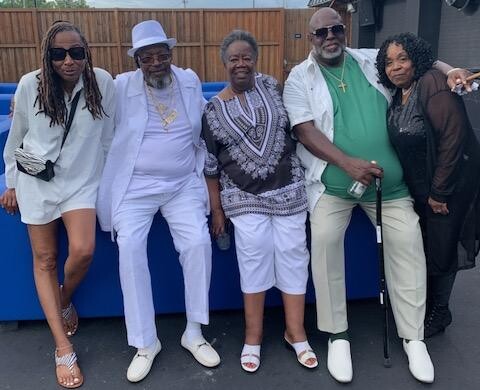 Five adults posing together outdoors, smiling for a group photo in casual and semi-formal summer outfits (left to right: woman in white dress with sunglasses, man in white suit and hat, woman in patterned top and white shorts, man in green shirt with white pants and cane, woman in dark top with cardigan).