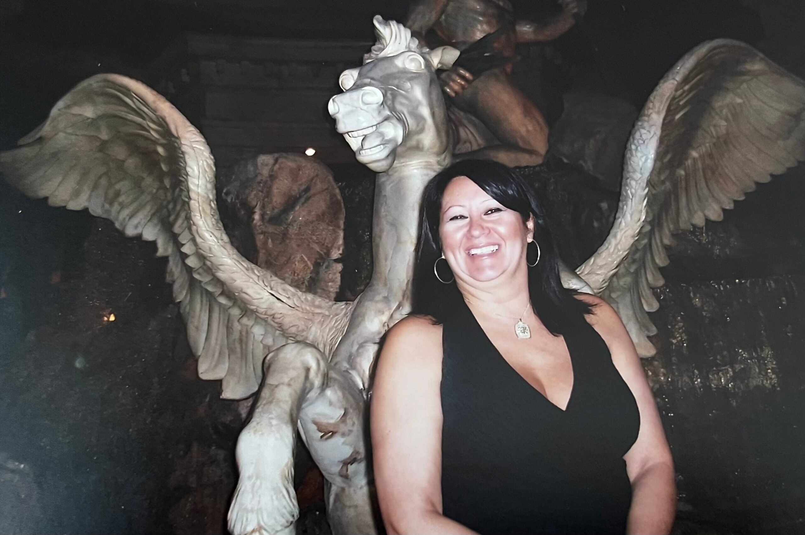 Smiling woman in a black dress posing beside a large stone statue with outstretched wings at night.