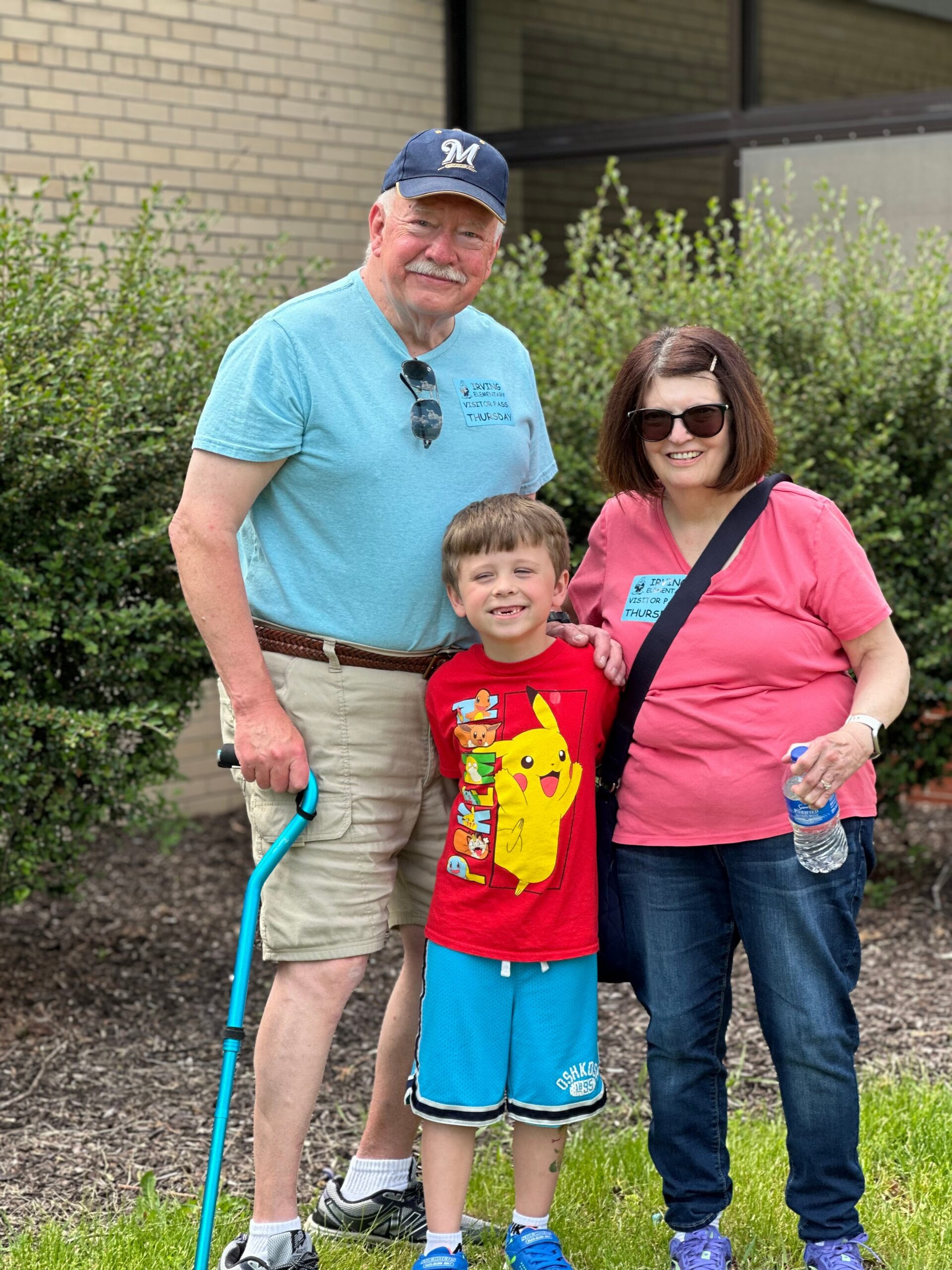 Smiling three‑person family pose outdoors: grandfather in a blue cap and turquoise shirt, mother in a pink shirt with sunglasses, and a boy in a red Pikachu shirt in front, with greenery and a brick building behind them.