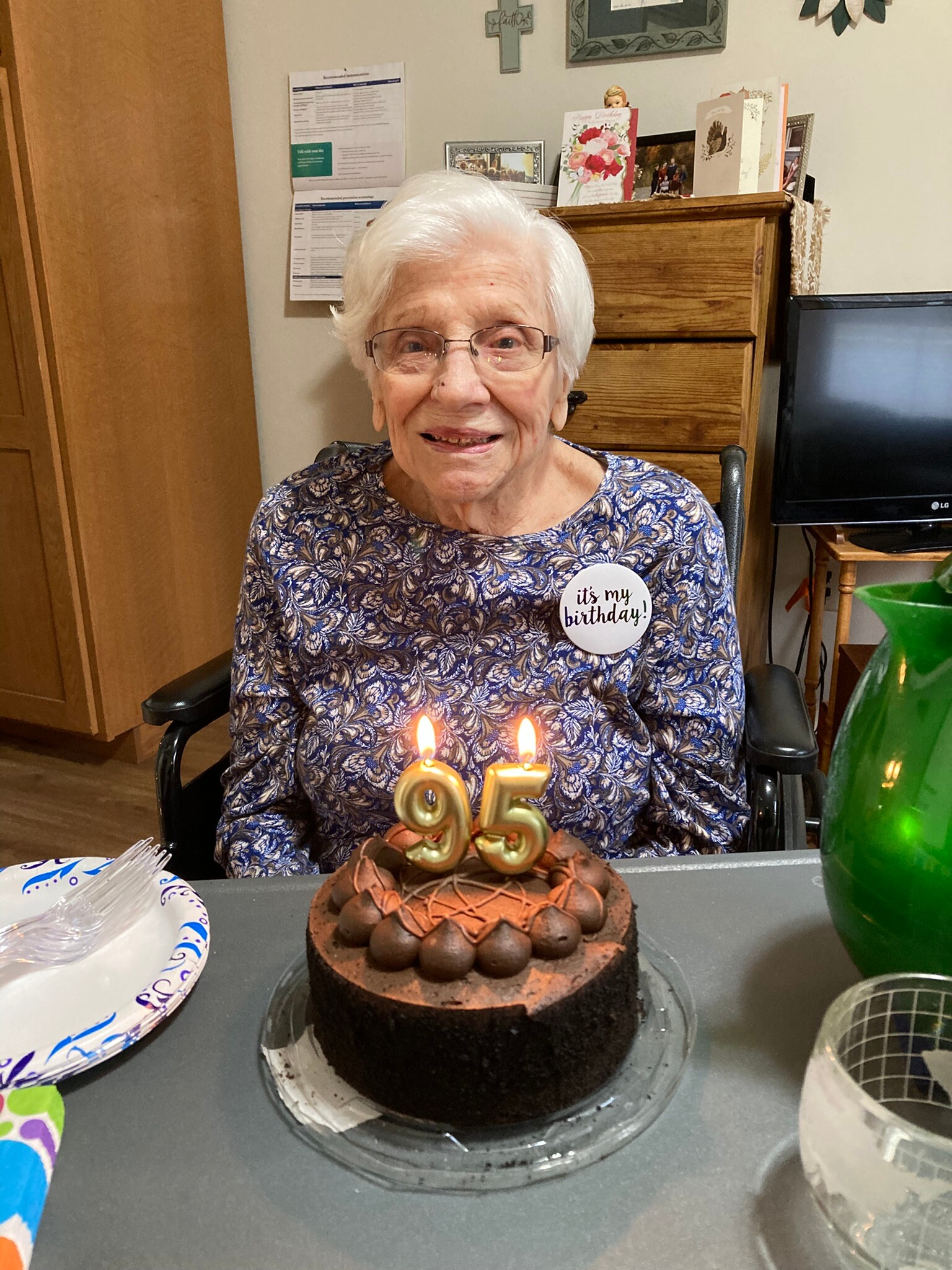 Smiling elderly woman in a wheelchair wearing a 'it's my birthday' badge, seated at a table with a chocolate cake and 95 candles in front of her.