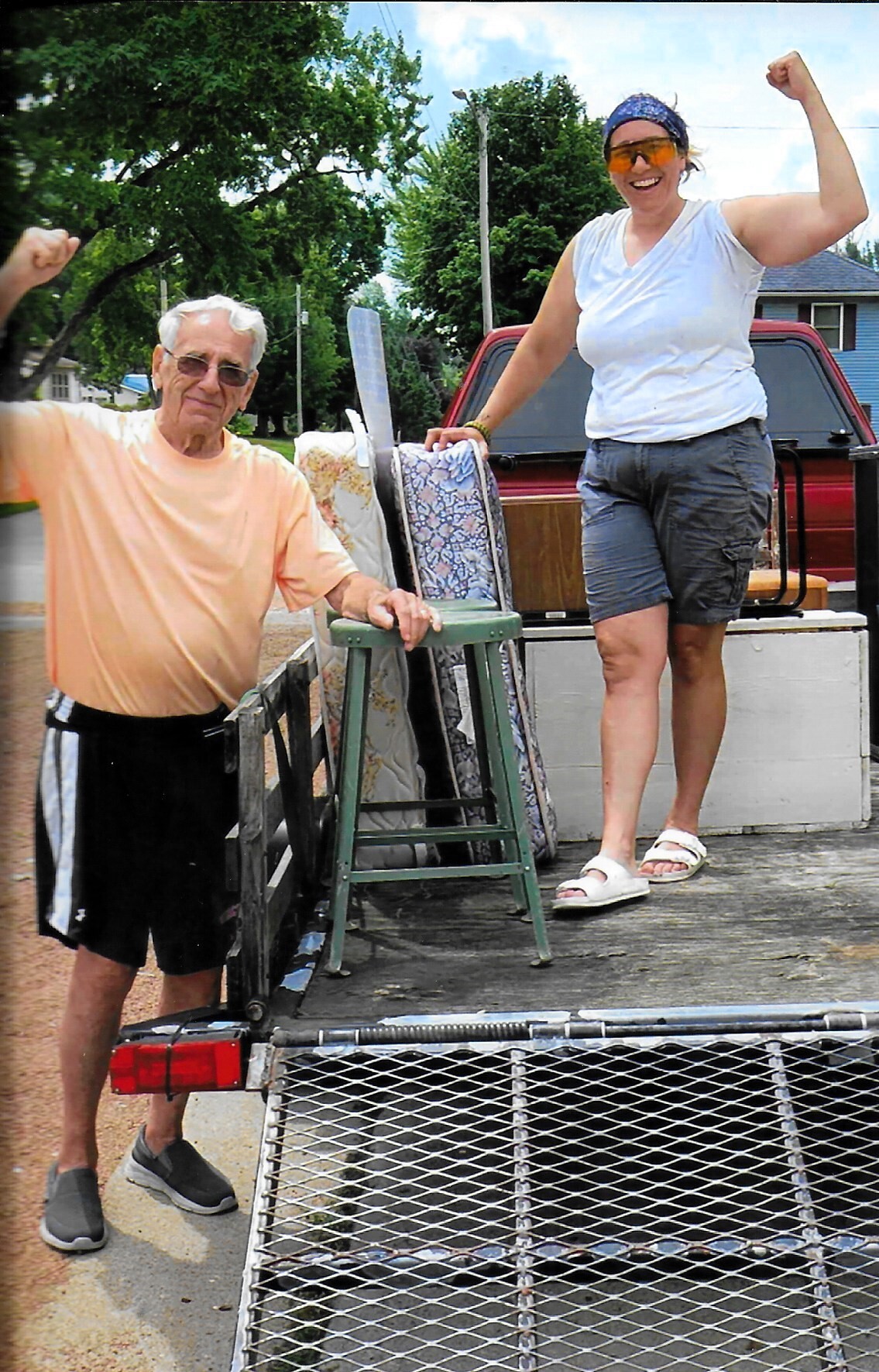 Two people stand on the back of a pickup truck moving household items, including a mattress and chairs, on a sunny day.