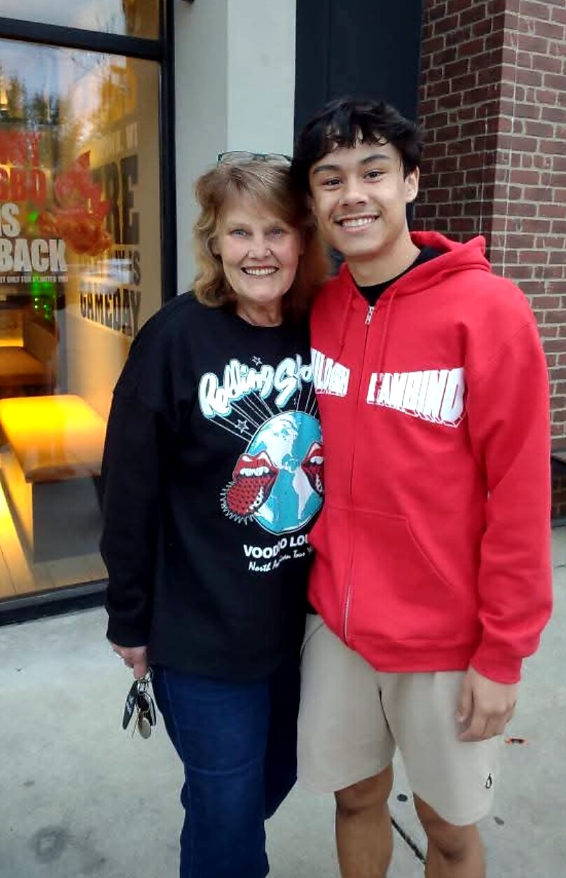 Smiling woman in a black graphic shirt stands next to a smiling young man in a red hoodie outside a brick storefront.