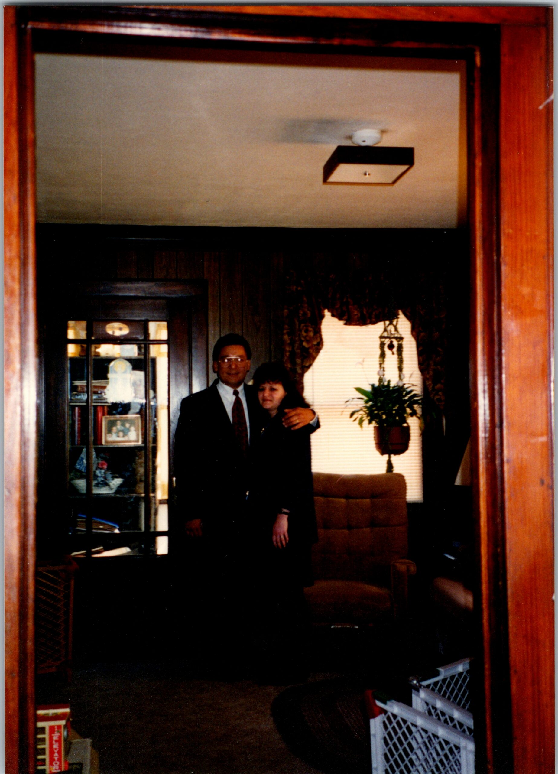 Man and woman in formal attire posing together in a wood-framed doorway, with a living room featuring a china cabinet and a potted plant behind them.