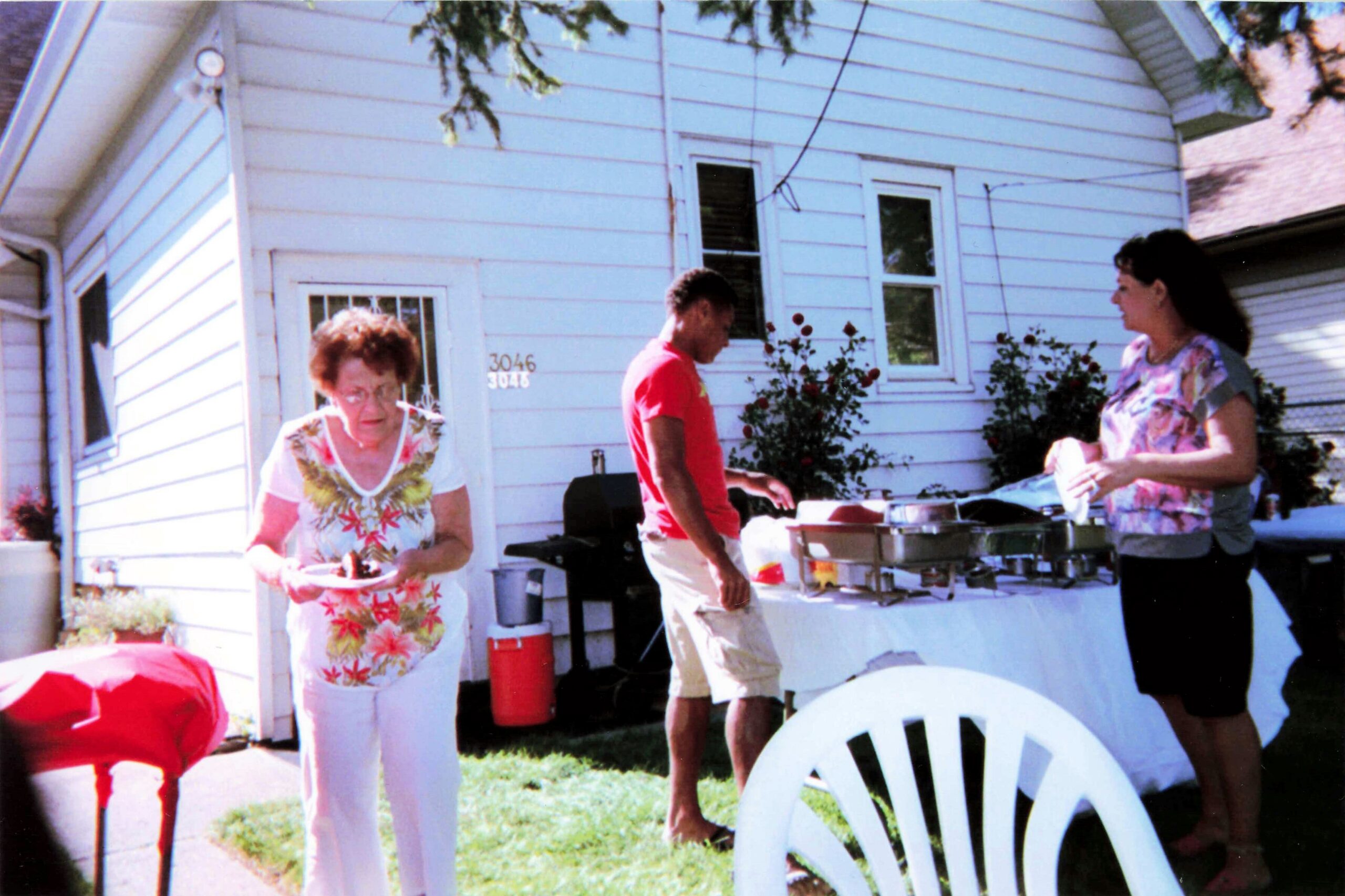 People at a backyard barbecue in front of a white house, with a woman in floral top serving food and a man in a red shirt nearby.