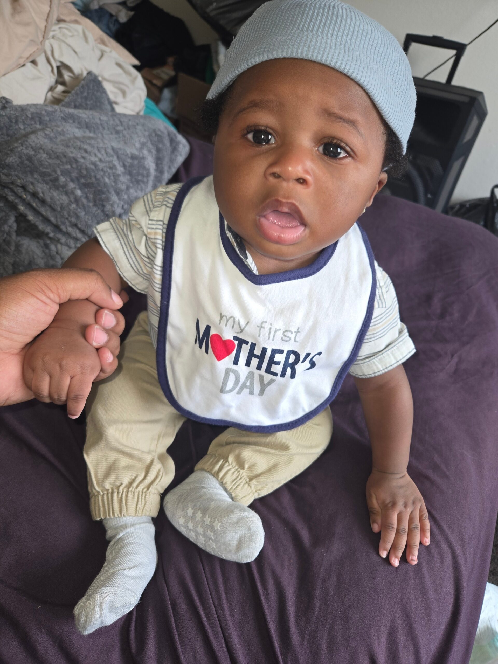 Baby with a light blue knit hat and a bib that reads 'My First Mother's Day,' holding an adult's finger on a dark purple couch.
