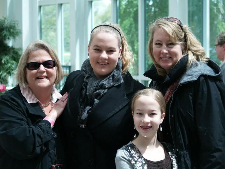Group portrait of four smiling people—two adult women, a teenage girl, and a younger girl—posing indoors with glass walls and greenery behind them.
