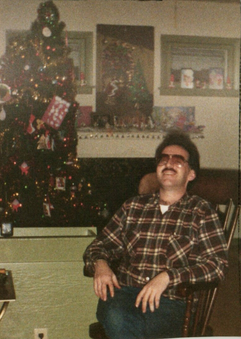 Man with a mustache wearing a plaid shirt sits in a chair beside a decorated Christmas tree.