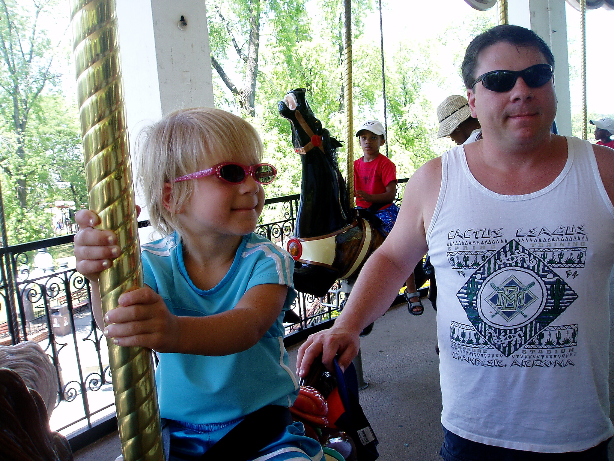 Young child in pink sunglasses grips a tall shiny gold pole on a carousel ride, with a man in sunglasses nearby.