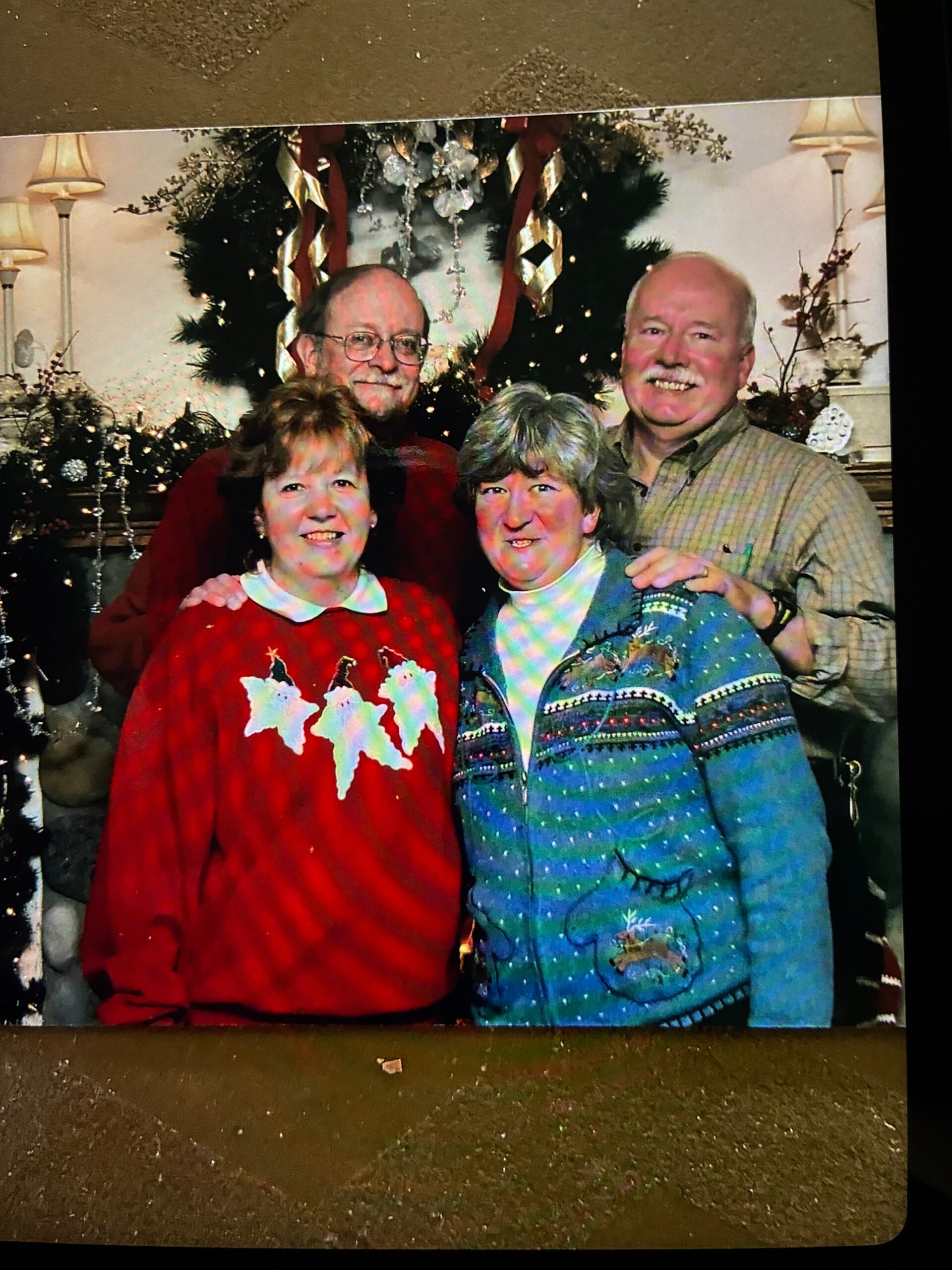 Four adults posing indoors in front of a decorated Christmas tree, wearing festive sweaters and smiling festive portrait.