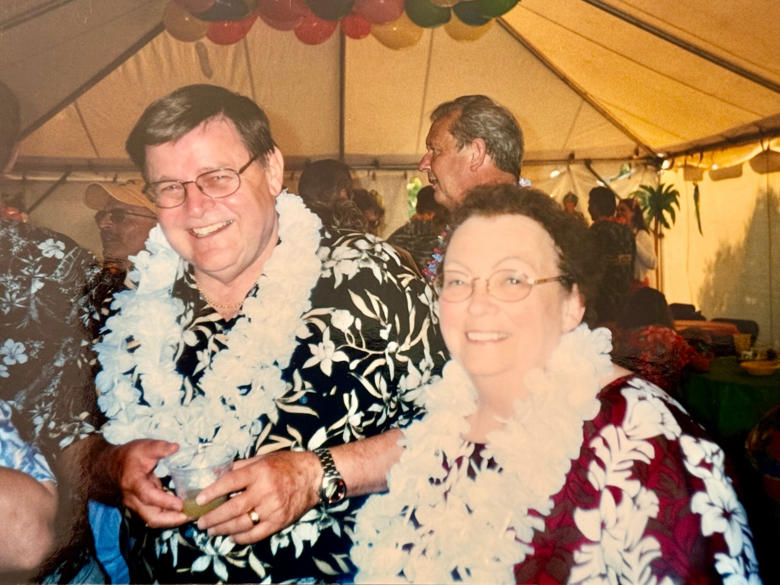 Two people in Hawaiian shirts with leis around their necks at a tropical-themed party under a tent, smiling for the camera.
