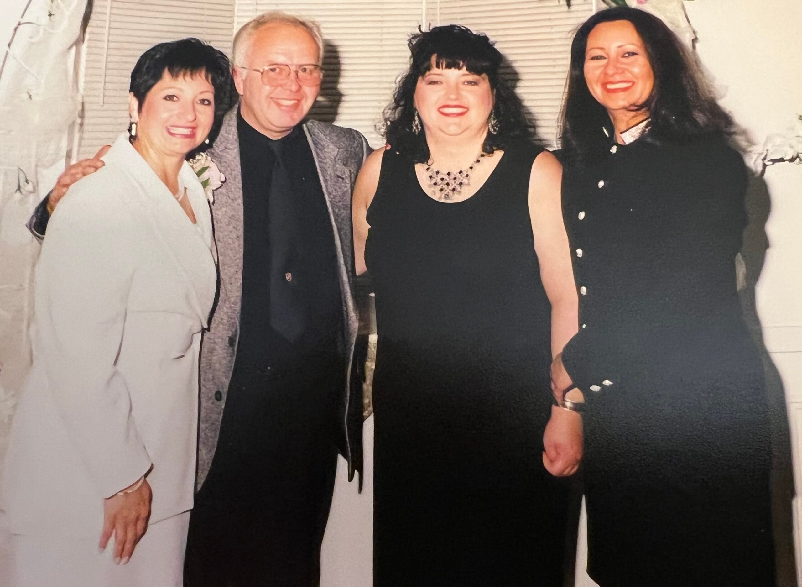 Group of four adults in formal outfits smiling for a photo at a celebration or event, standing close together indoors.