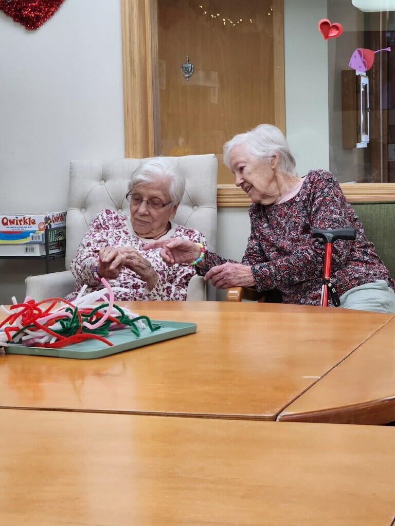 Two elderly women sit at a wooden table, weaving colorful cords into a craft project, with a walker nearby.