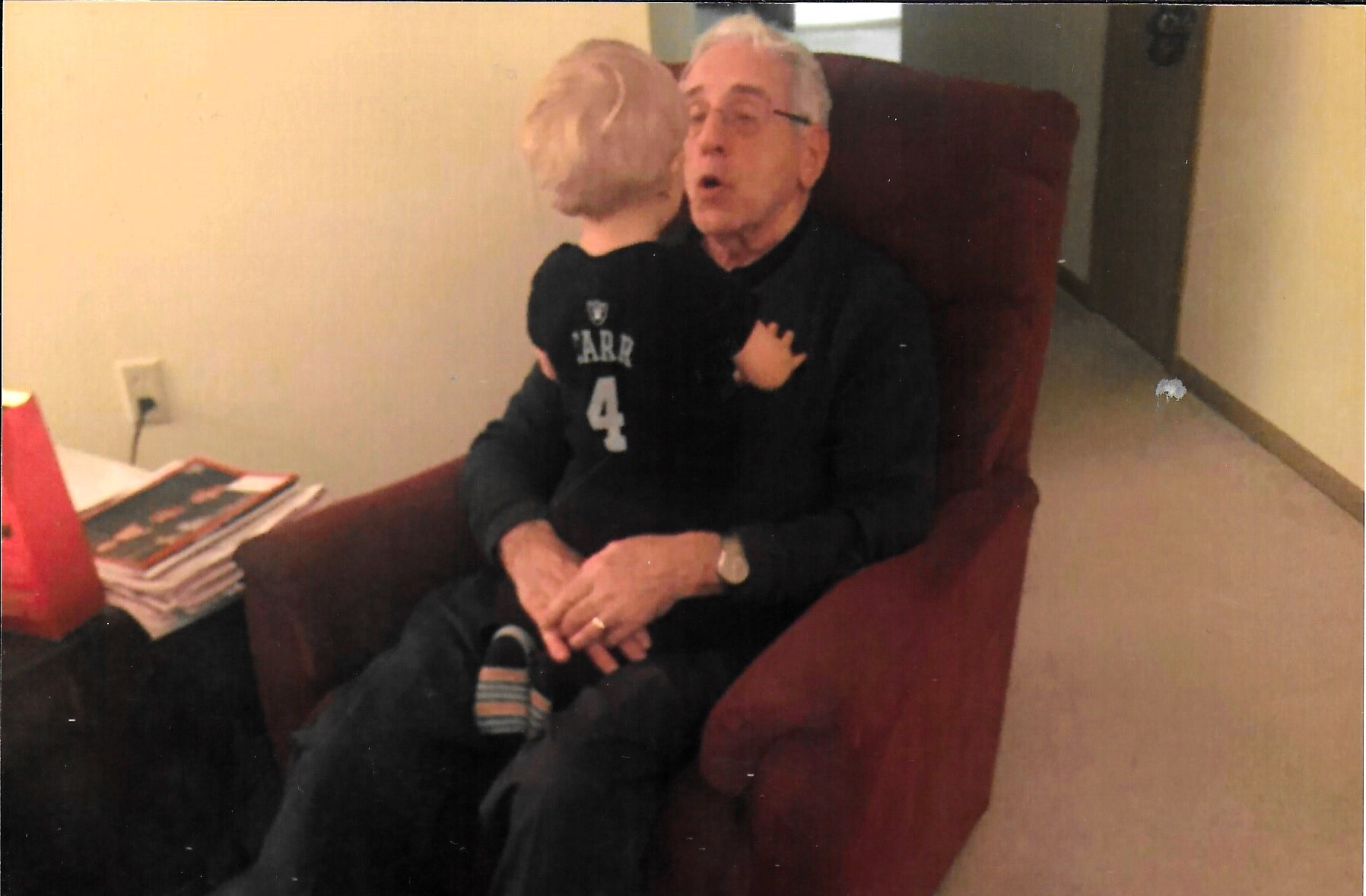An elderly man with glasses sits in a red chair while a child kisses his cheek; a stack of magazines sits on a nearby table.