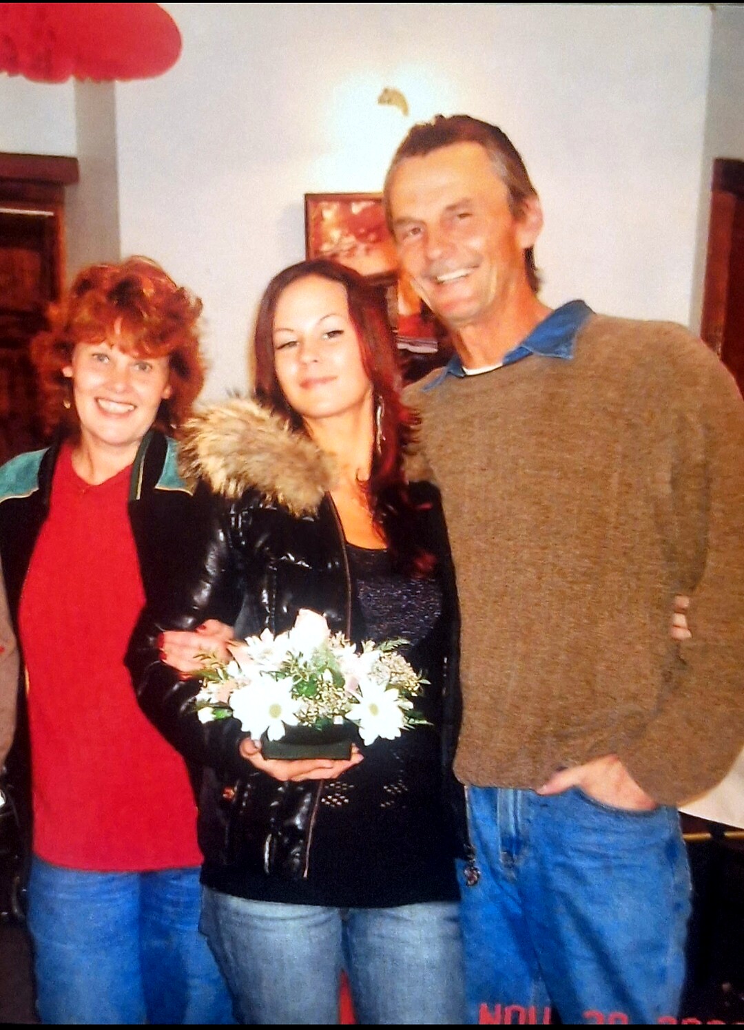Three people posing together indoors; woman center holds a small bouquet of white flowers and wears a black jacket. Left woman smiles, right man leans in.