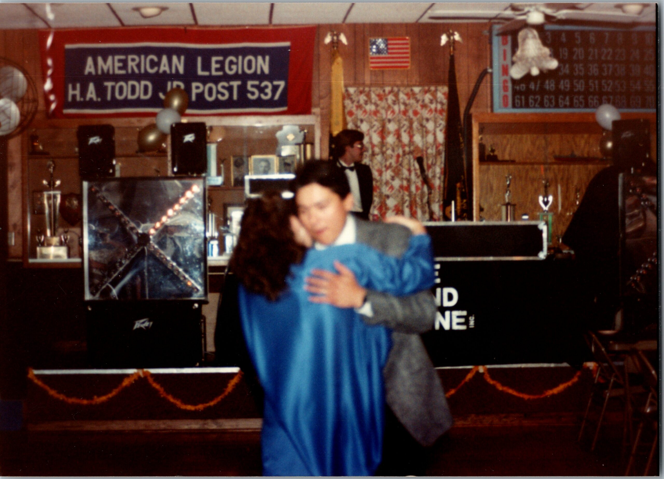 Couple dancing in a wood-paneled hall with an American Legion banner reading 'American Legion H.A. Todd Jr Post 537' in the background.