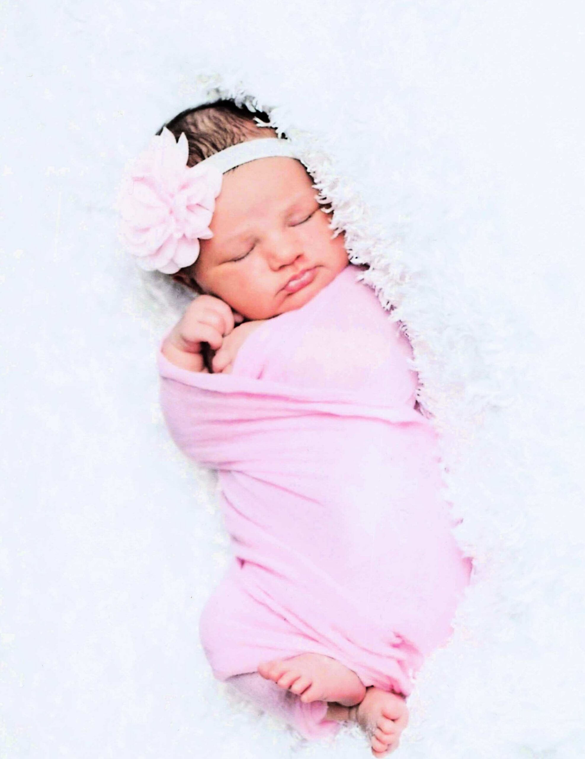 Newborn baby girl peacefully sleeping, wrapped in a pink blanket with a pink flower headband on a soft white background
