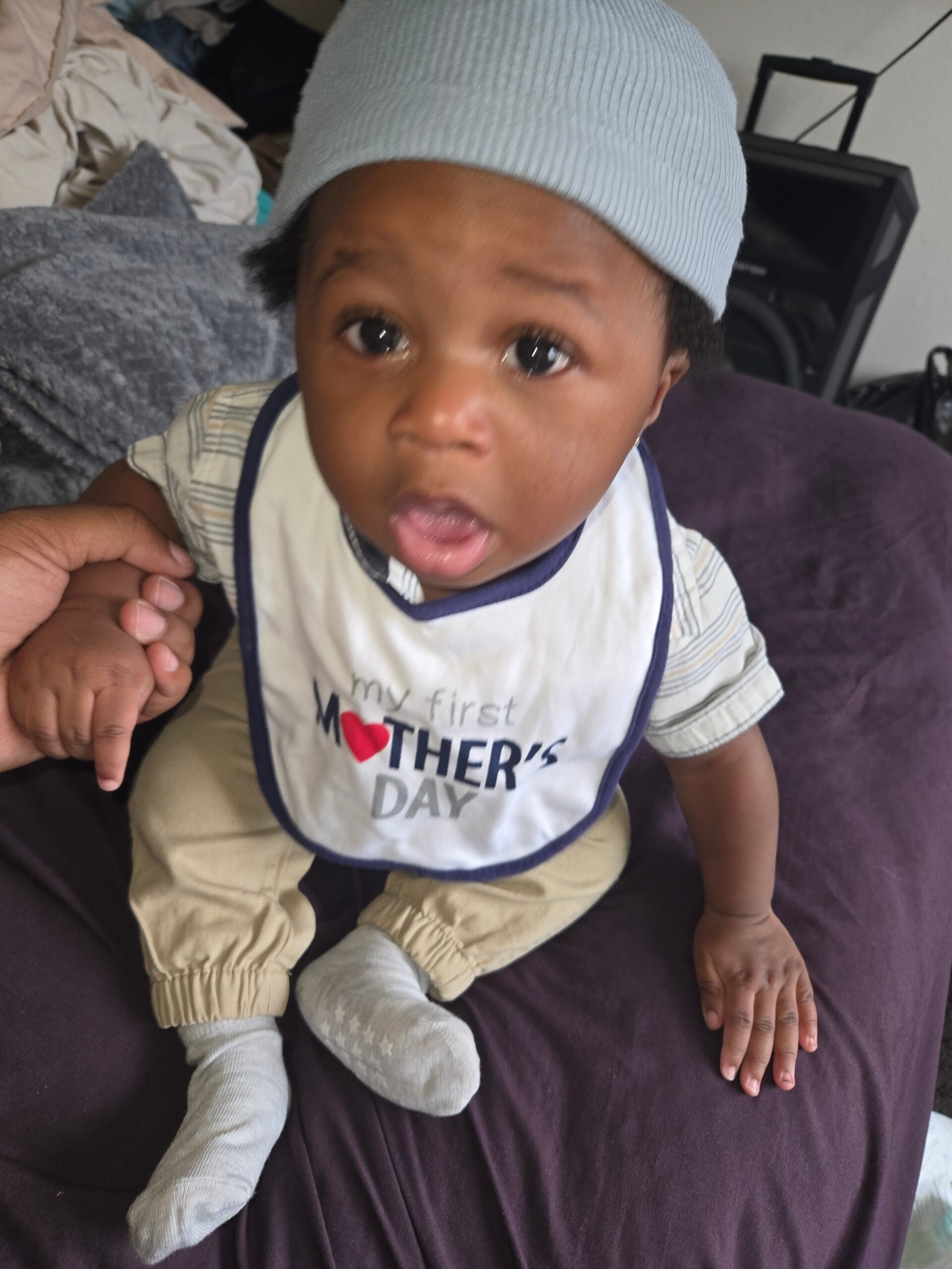 Close-up of a baby wearing a light blue knit cap and striped shirt, with a bib that reads 