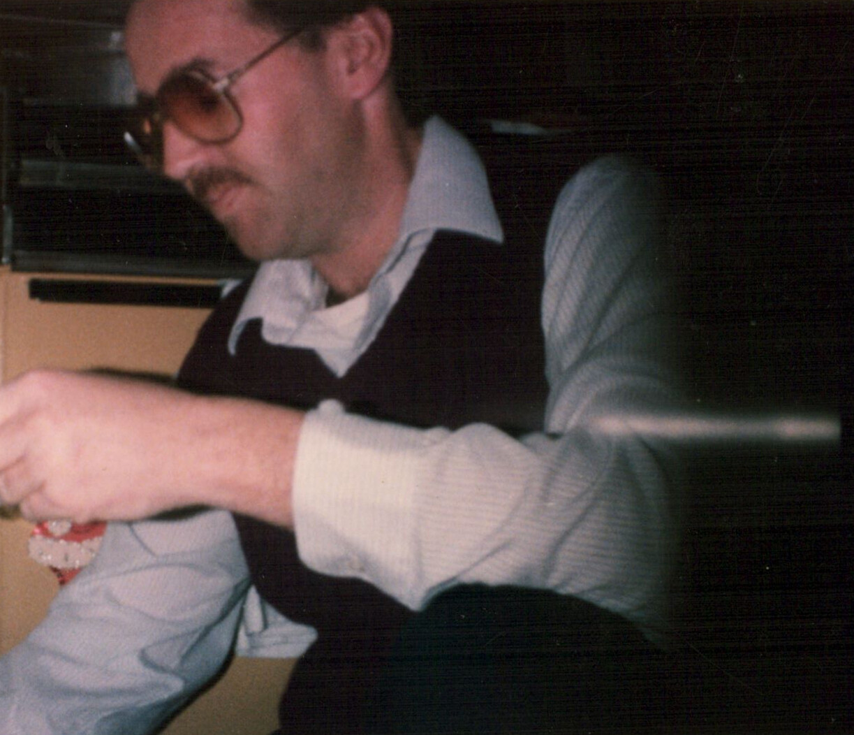 Man with a mustache and sunglasses, wearing a light blue shirt and dark vest, looking down while handling a small object.