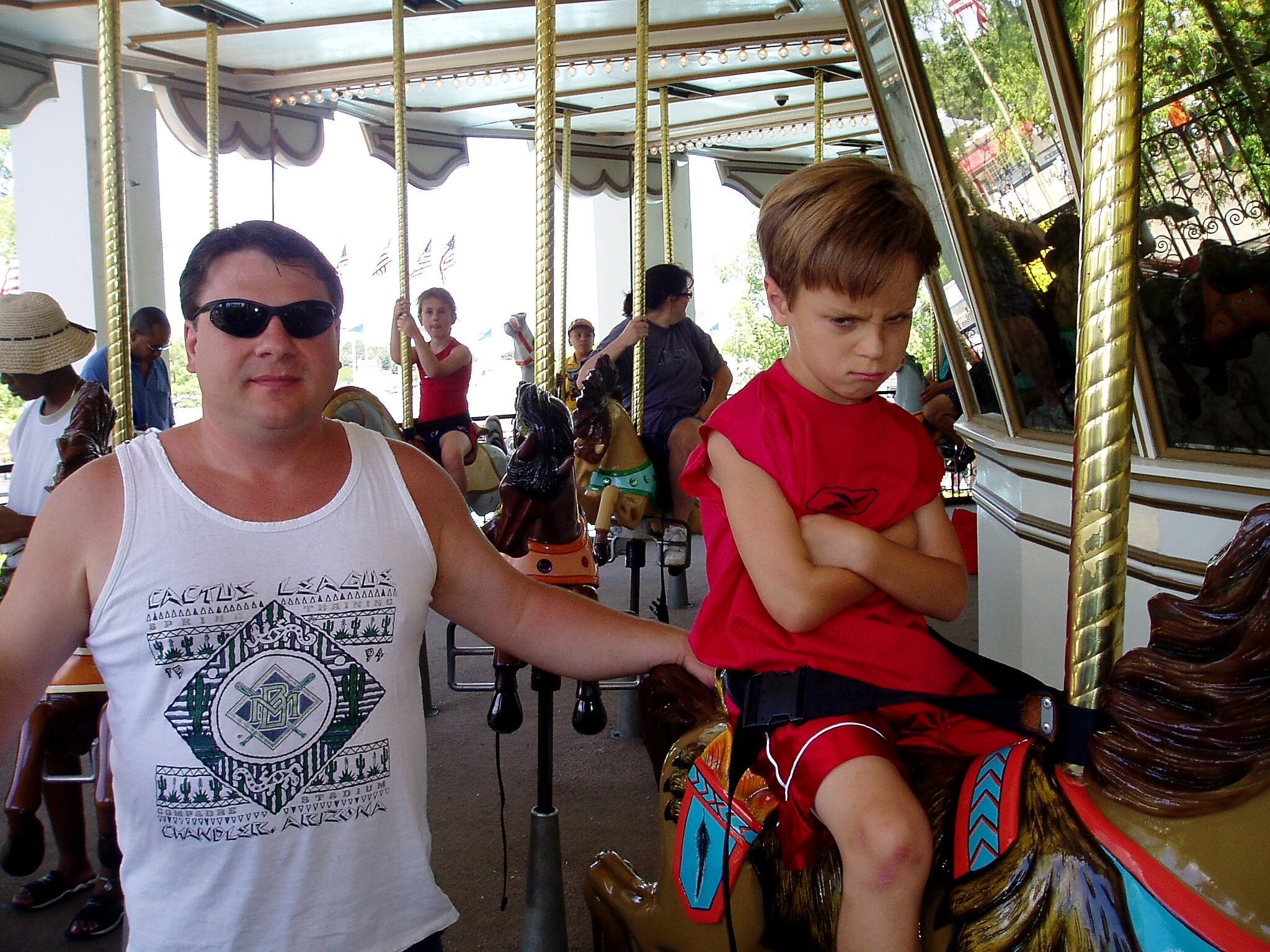 A man in a white tank top and sunglasses sits on a carousel next to a red-shirted child looking down, with gold poles and decorative horses around them.