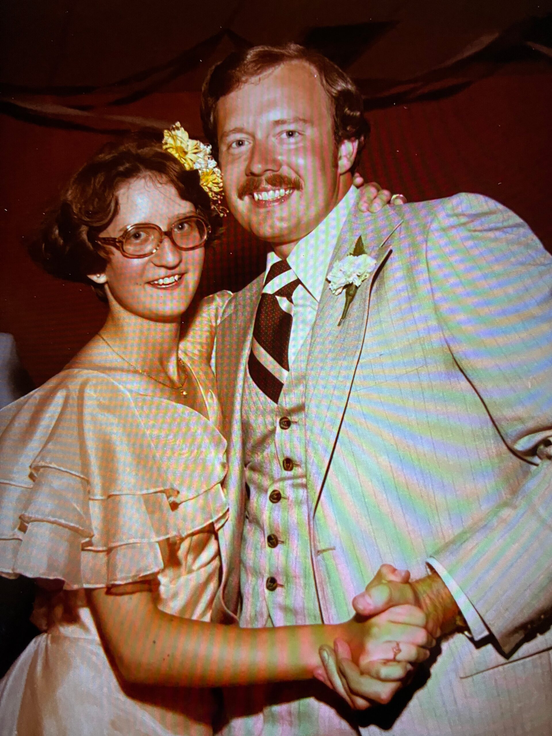 Smiling couple dancing at a wedding: woman with short hair, glasses, and a yellow flower in her hair; man in a light suit with a striped tie and boutonniere.