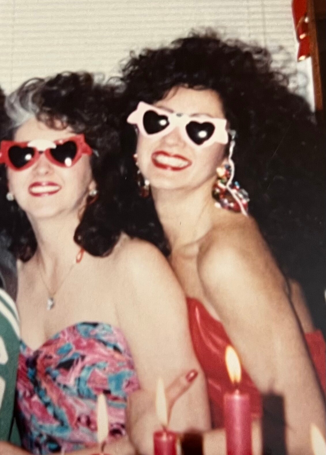 Two women in colorful outfits wearing heart-shaped sunglasses at a party, smiling beside lit candles on the table.