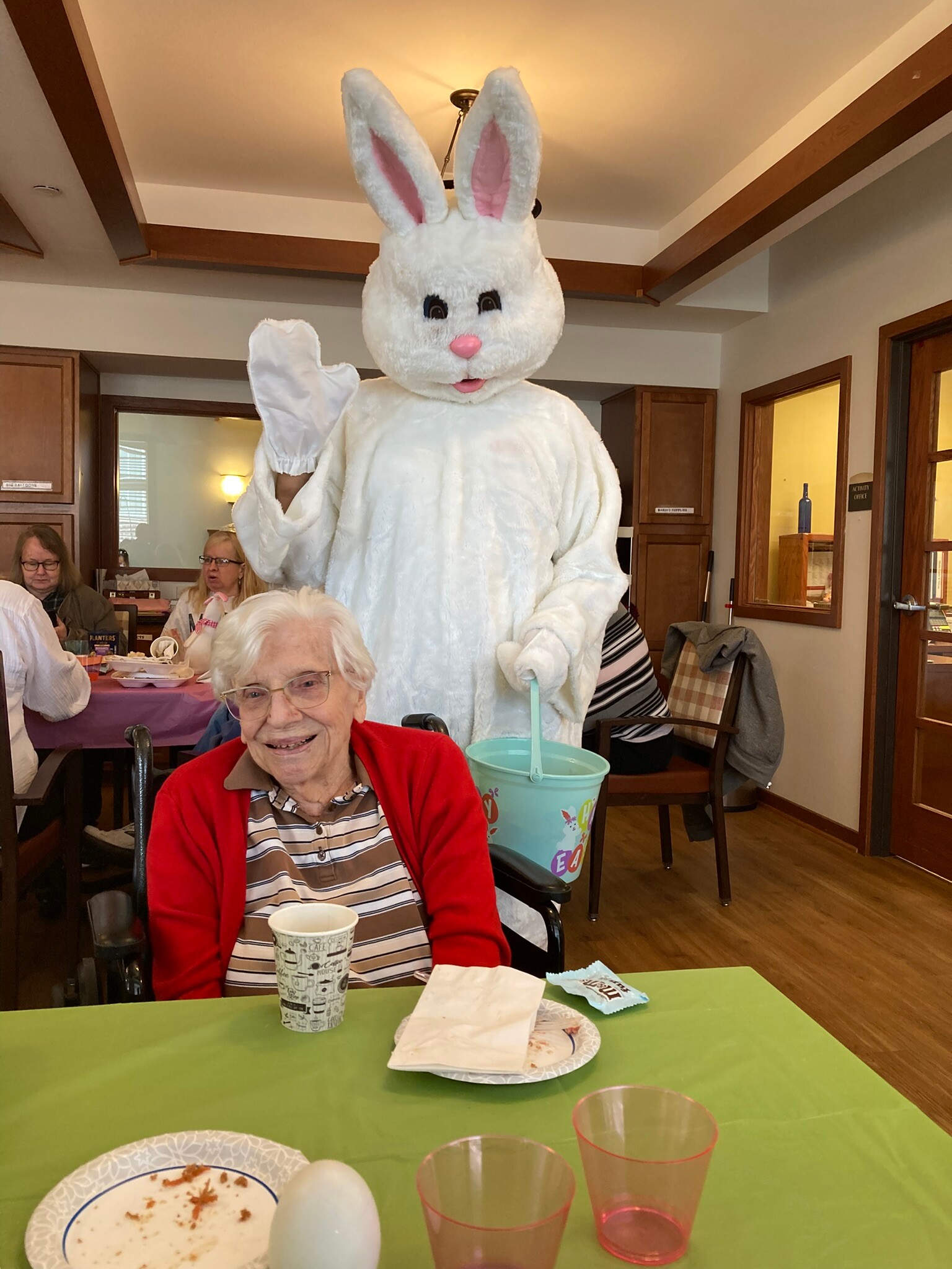 Person in a large white Easter Bunny costume waving at a table of seniors in a communal dining room during a celebration.