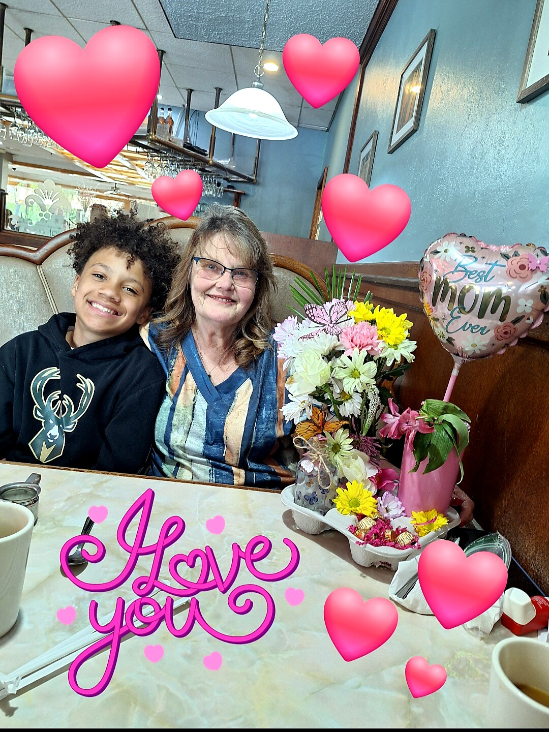 Smiling boy and woman sit at a restaurant booth with a flower bouquet and a pink 'Best Mom Ever' balloon, pink heart overlays, and 'I Love You' text sticker.
