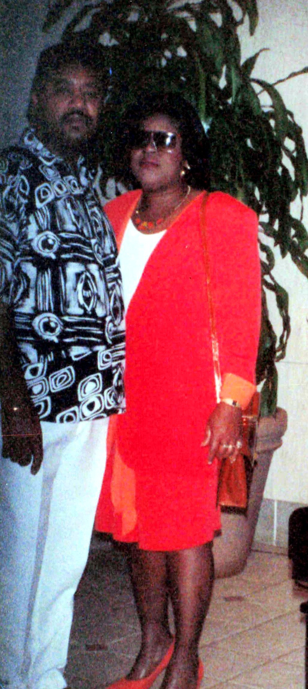 Woman in a bright orange-red coat over a white top, wearing sunglasses, standing next to a man in a black-and-white patterned shirt indoors near a tall plant.