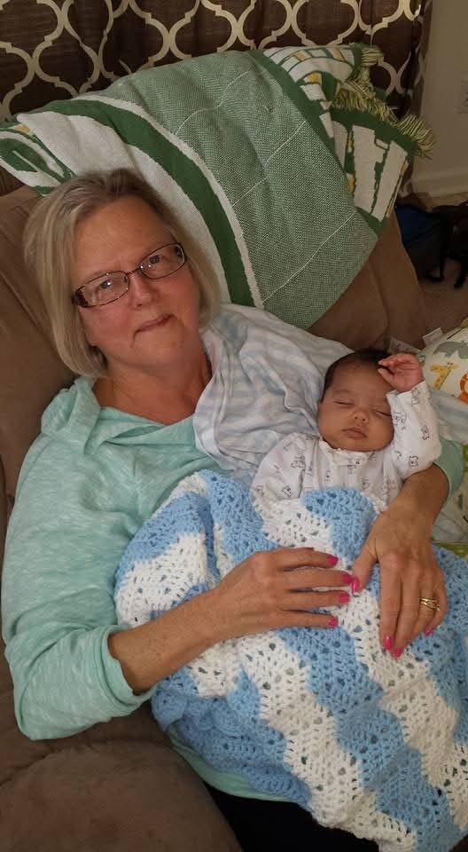 Older woman with glasses resting on a couch, cradling a sleeping baby wrapped in a blue-and-white crochet blanket.