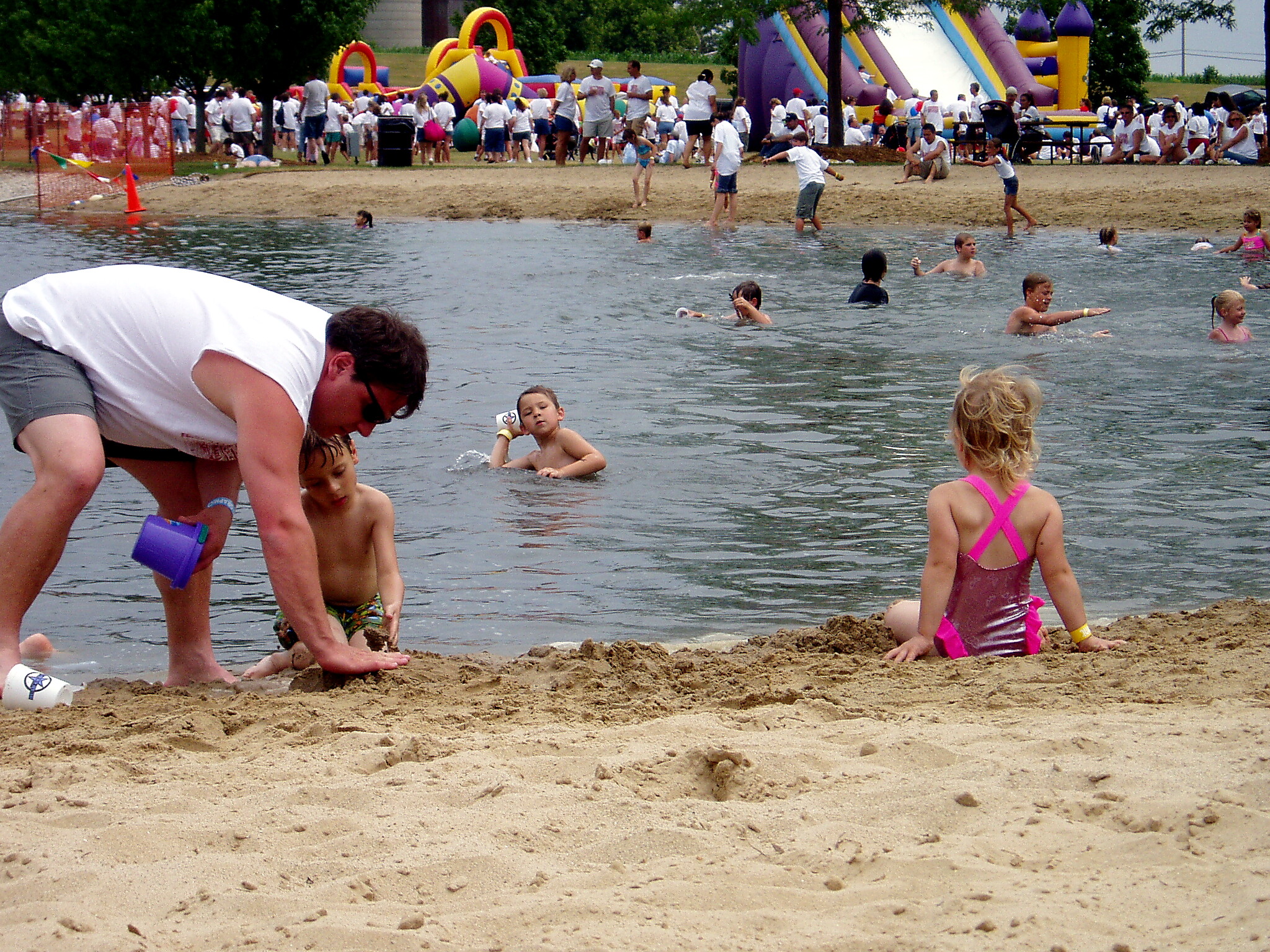 Families and kids enjoy a lakeside beach with sand, water play, and colorful inflatable obstacle course in the background.