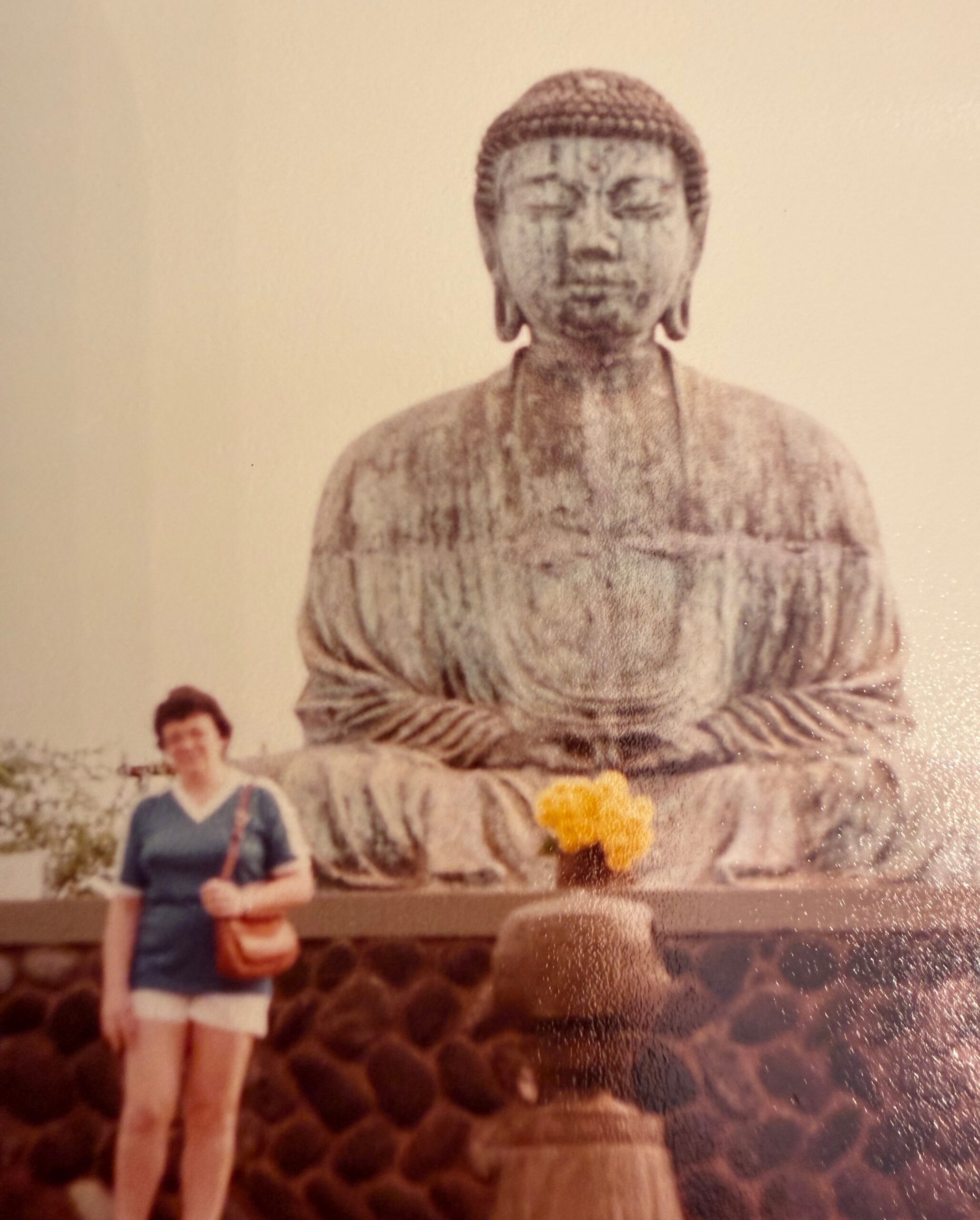 Large stone Buddha statue sitting with hands resting in lap, foreground tourists in casual clothing at a rocky platform.