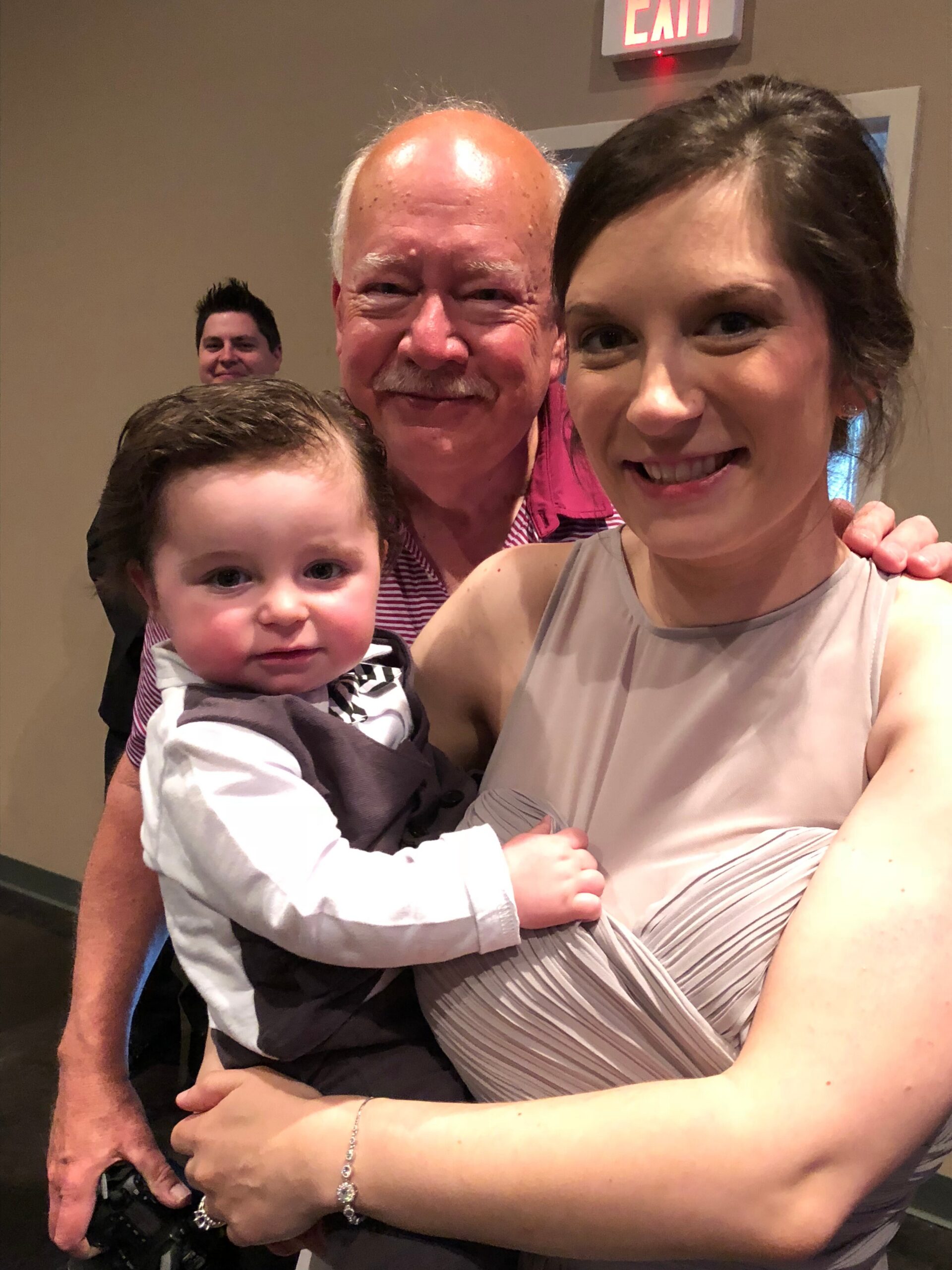 Woman in a sleeveless dress smiling, holding a baby; a man stands behind them, exit sign in the background.