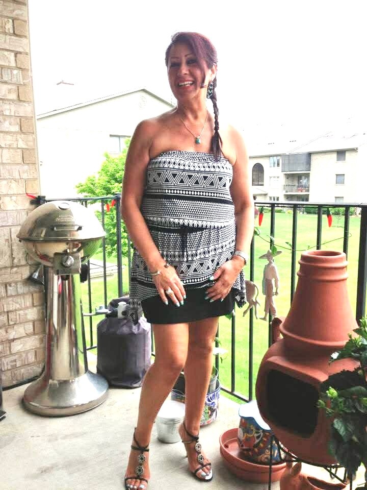 Smiling woman in a black-and-white geometric strapless dress and sandals posing on a balcony with a grill nearby.