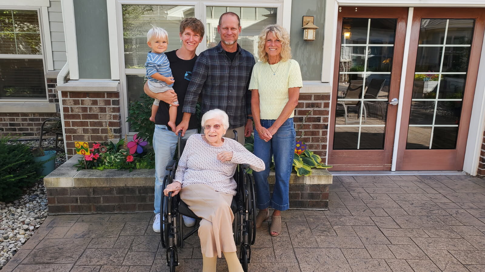 Grandmother in a wheelchair with family posing on a brick-house porch, colorful flowers nearby.