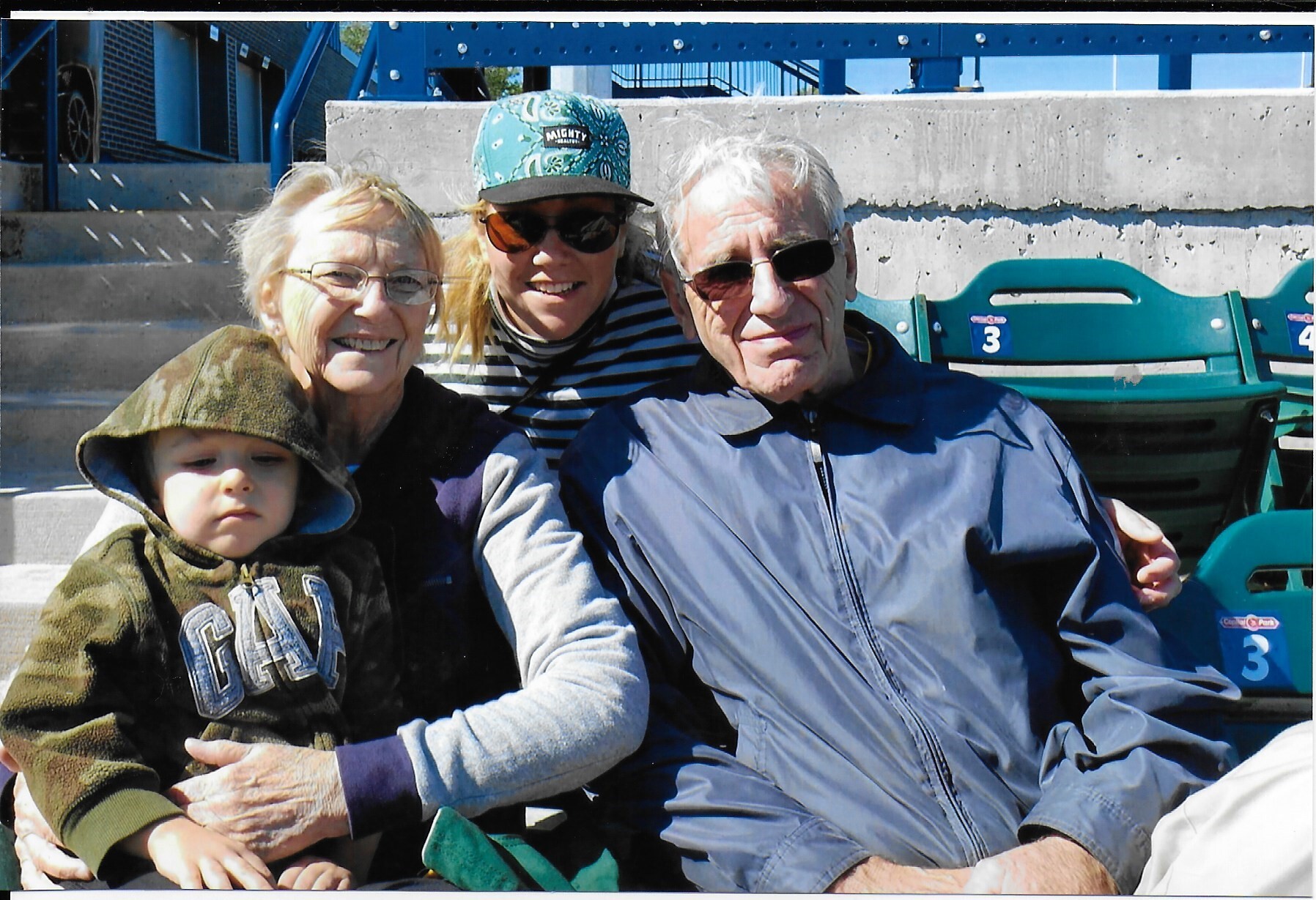 Smiling elderly woman, man, and a younger woman with a cap pose together with a small child on blue stadium bleachers outdoors.